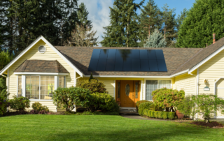 Yellow house with solar panels on the roof, surrounded by lush greenery and a well-maintained lawn.