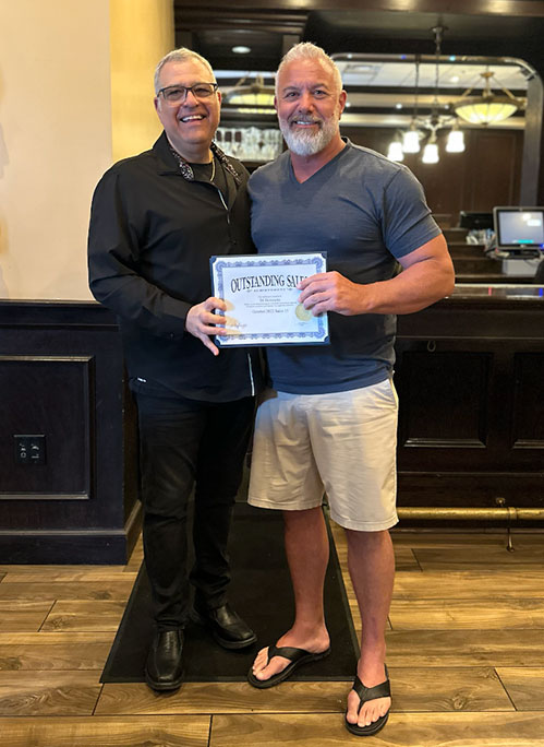 Photo of Al and award recipient smiling, holding a certificate of Outstanding Sales, standing in a restaurant or bar setting.