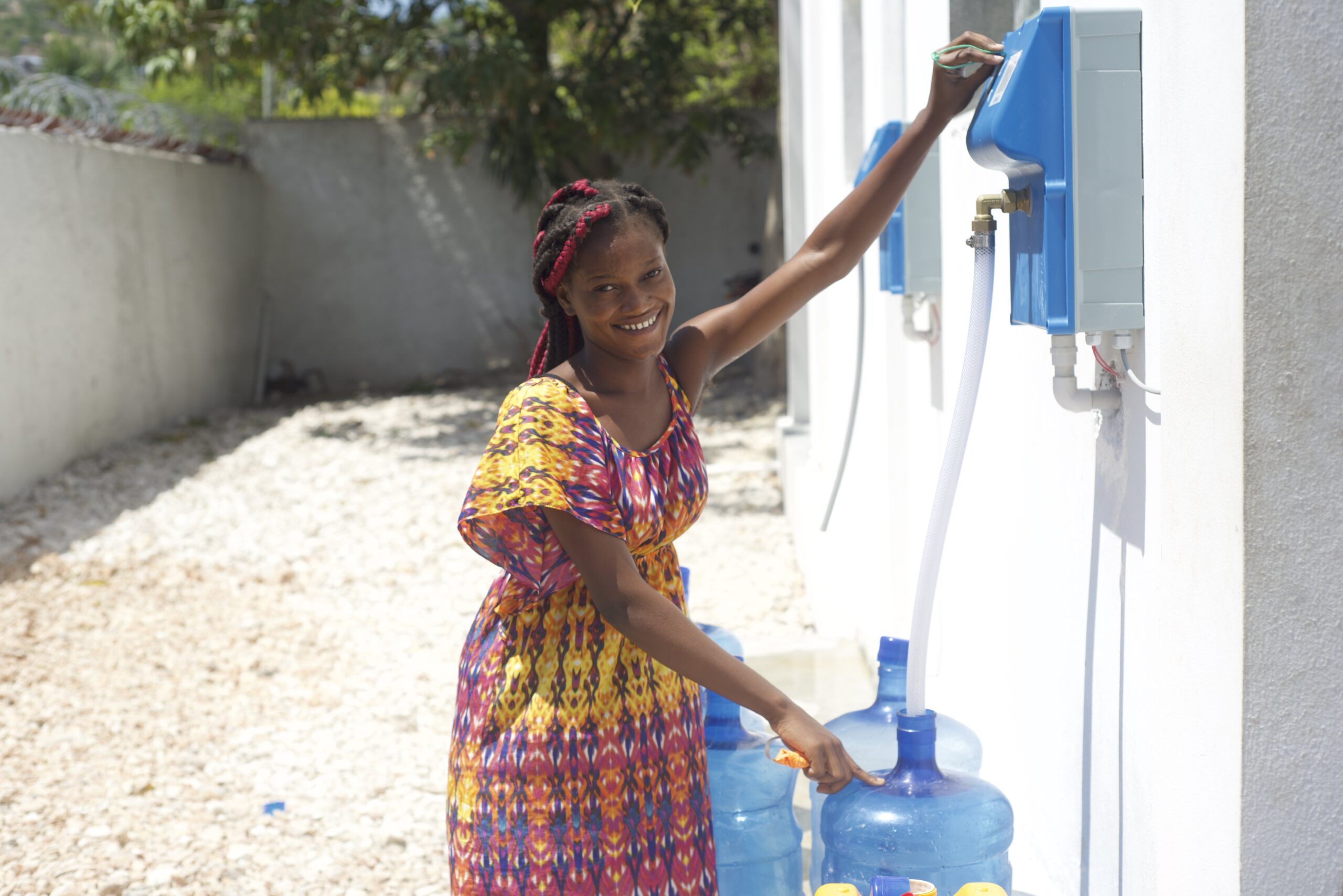 Smiling woman in a colorful dress filling a water container from an outdoor wall-mounted dispenser.
