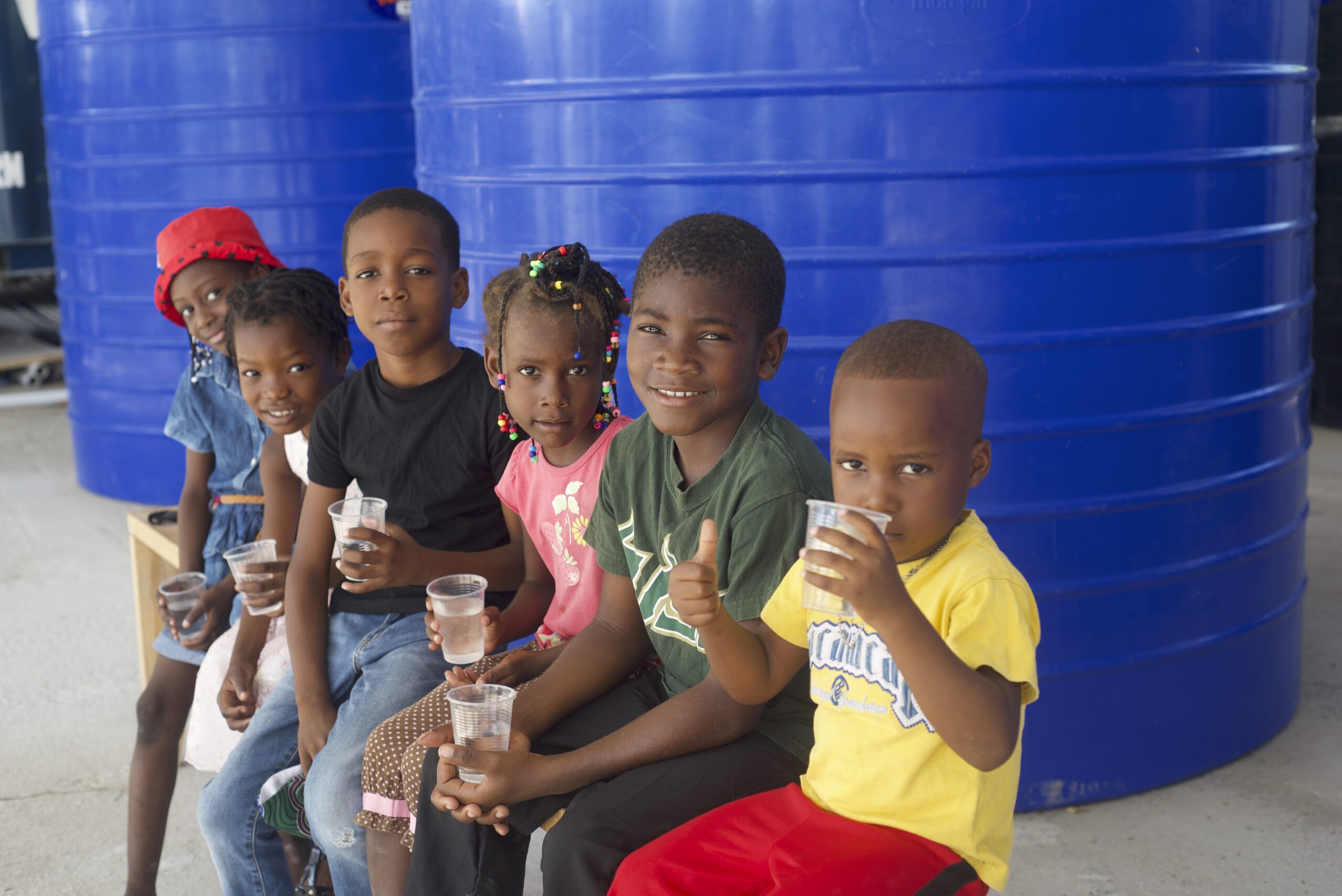 Group of children sitting in front of large water tanks, holding cups and smiling.